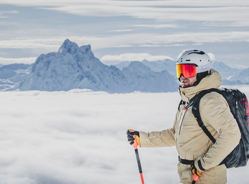 Ein Skifahrer steht mit Stock und Rucksack auf einem schneebedeckten Gipfel über einer Wolkendecke mit Bergen im Hintergrund.