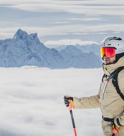 Ein Skifahrer steht mit Stock und Rucksack auf einem schneebedeckten Gipfel über einer Wolkendecke mit Bergen im Hintergrund.