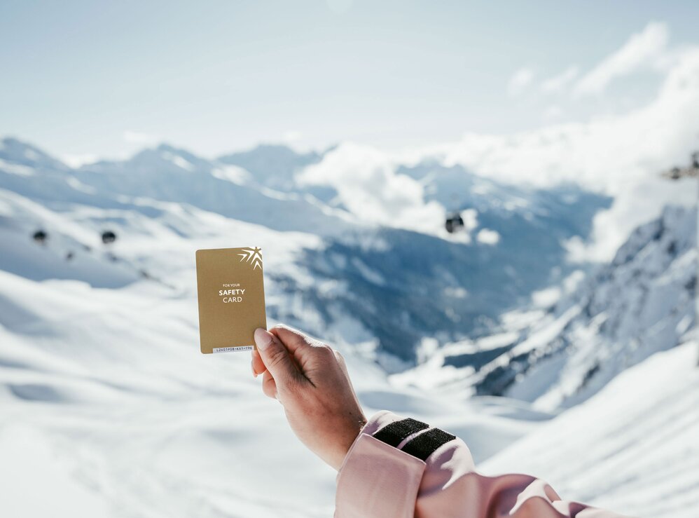 Person hält einen Skipass vor schneebedeckten Alpen.