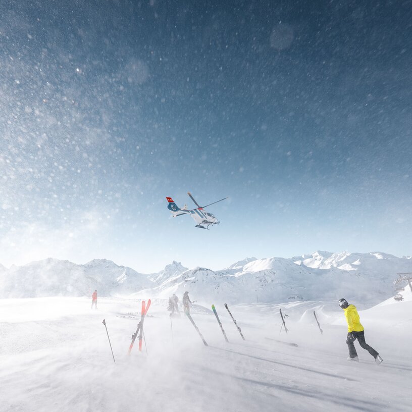 Helikopterlandung auf verschneiter Skipiste mit Skifahrer und Bergbanorama im Hintergrund Schnee wirbelt durch die Luft.