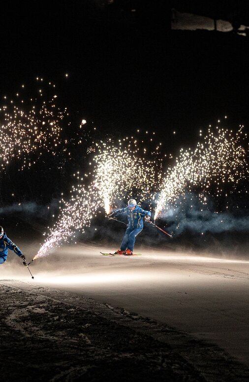 Zwei Skifahrer fahren nachts mit funkensprühenden Fackeln.