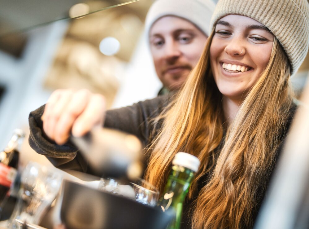 Ein lachendes Paar holt sich am Buffett auf einer Skihütte etwas zu essen und zu trinken. Sie tragen warme Kleidung und Mützen.