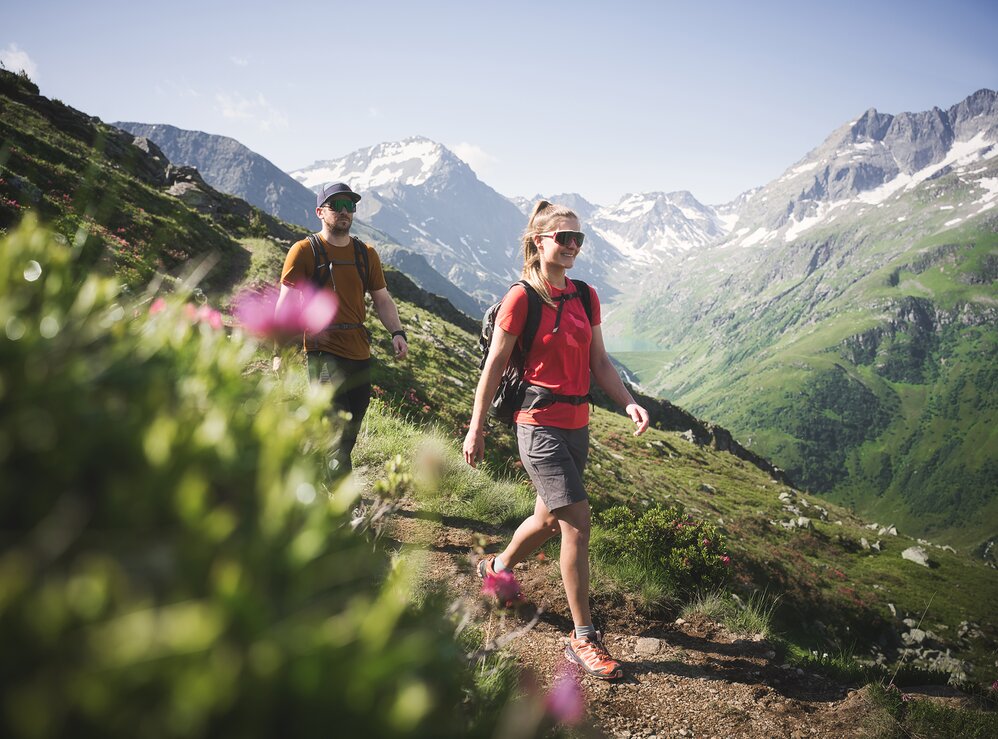 Zwei Personen wandern durch die grüne Natur in den Bergen von St. Anton am Arlberg mit schneebedeckten Gipfeln im Hintergrund.
