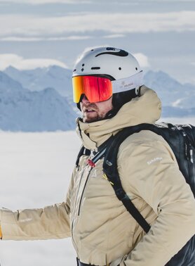 Ein Skifahrer mit Helm und Rucksack über den Wolken vor schneebedeckten Bergen.
