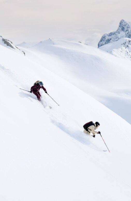 Zwei Skifahrer gleiten dynamisch durch unberührten Pulverschnee vor einer beeindruckenden Bergkulisse.