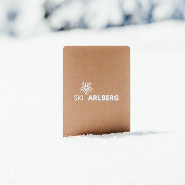 Ein goldener Skipass von Ski Arlberg steckt im Schnee, im Hintergrund verschneite Berggipfel.