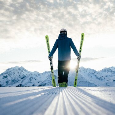 Eine Person mit Skiern vor einer malerischen Berglandschaft im Sonnenschein.