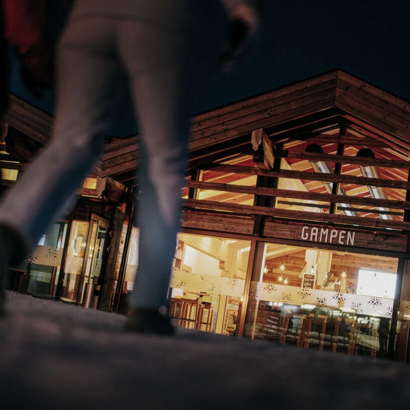 Zwei Personen gehen bei Nacht auf das beleuchtete Gampen Restaurant zu.