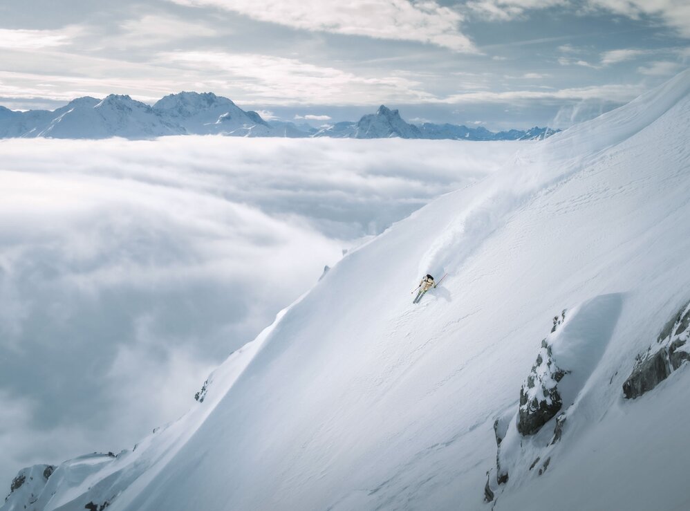 Ein Skifahrer fährt eine steile Piste in der Skiregion St. Anton am Arlberg hinunter, umgeben von Wolken und Berggipfeln.
