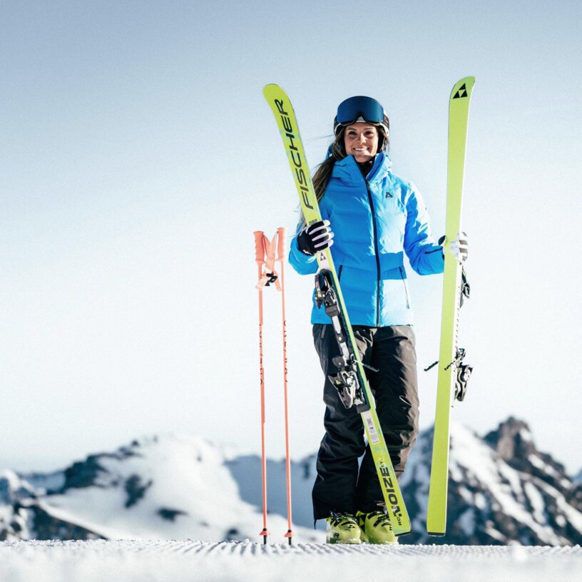 Eine Frau steht auf einer Piste mit Skiern und Helm, Berge im Hintergrund.
