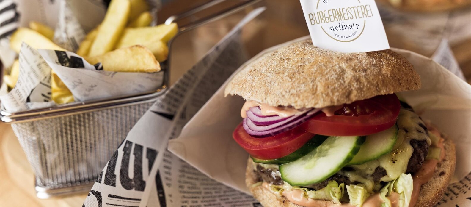Ein vegetarischer Burger mit roter Zwiebel, Tomate und Gurke, serviert mit Pommes in einem Metallkorb auf Zeitungspapier.