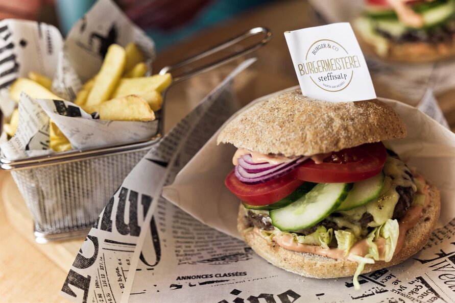 Ein vegetarischer Burger mit roter Zwiebel, Tomate und Gurke, serviert mit Pommes in einem Metallkorb auf Zeitungspapier.