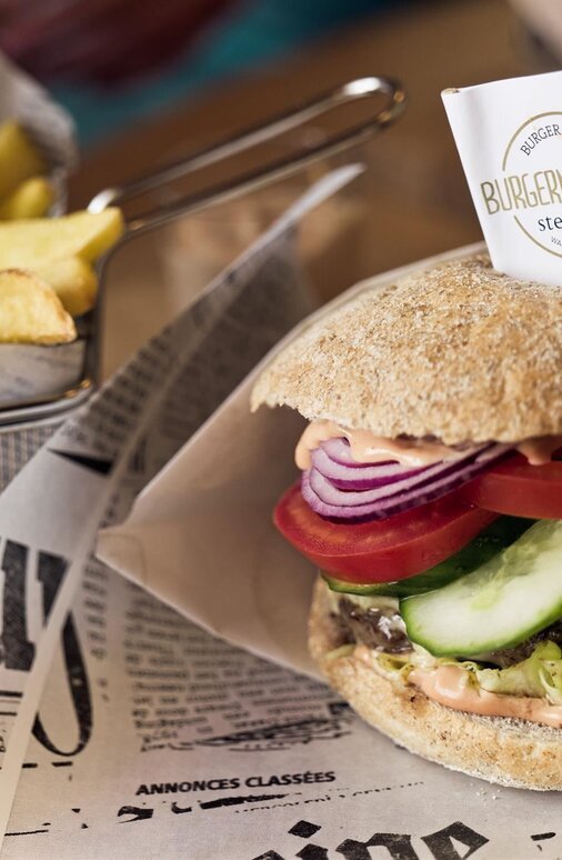 Ein vegetarischer Burger mit roter Zwiebel, Tomate und Gurke, serviert mit Pommes in einem Metallkorb auf Zeitungspapier.