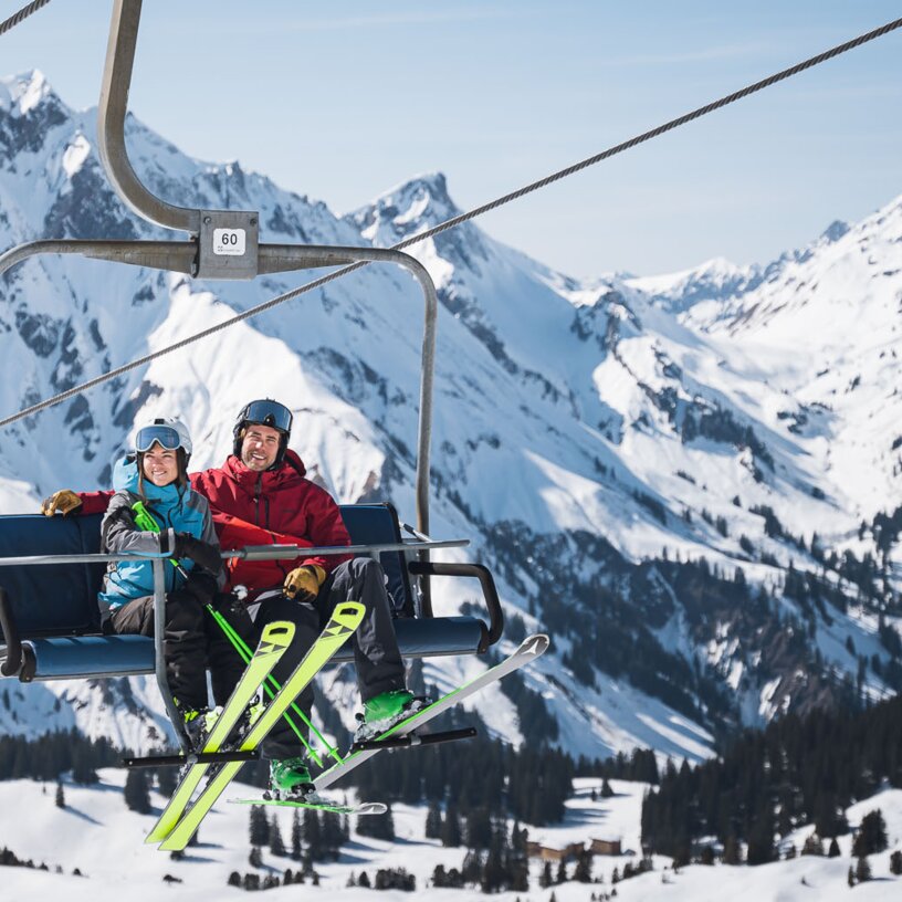 Zwei Personen sitzen auf einem Sessellift in einer schneebedeckten Berglandschaft.
