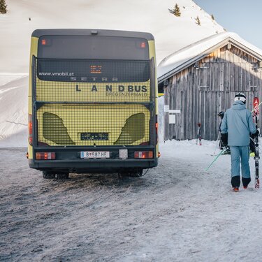 Zwei Skifahrer gehen von einem Bus zur Piste. Der Bus trägt die Aufschrift 'vmobil.at' und 'Landbus Bregenzerwald'.