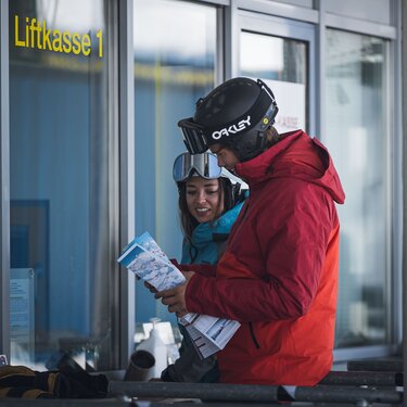 Zwei Menschen im Skianzug vor der Liftkasse mit einer Karte in der Hand.