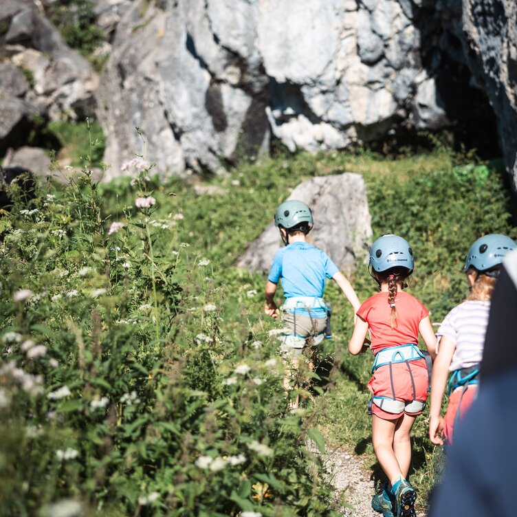 Kinder mit Helmen und Klettergurten auf einem schmalen Pfad im Freien.