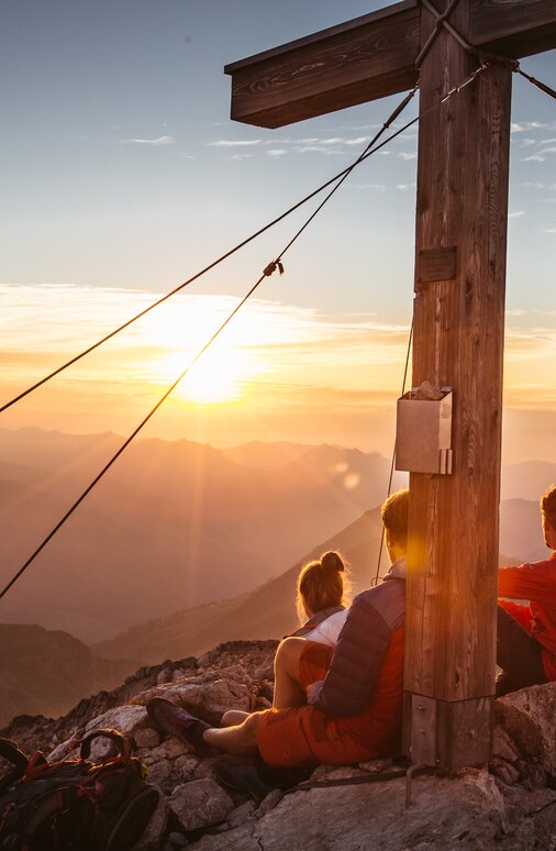 Zwei Personen sitzen bei Sonnenaufgang auf einem Berg neben einem Gipfelkreuz.