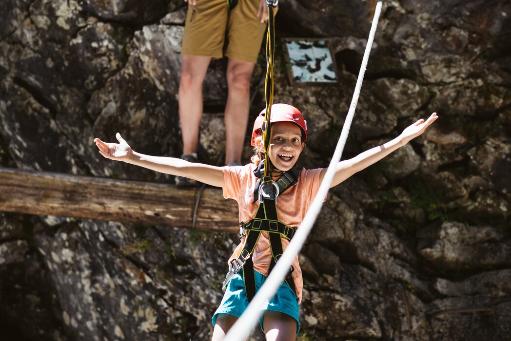 Kind in Sicherheitsausrüstung fährt mit ausgebreiteten Armen über die Zipline, eine erwachsene Begleitperson steht im Hintergrund.