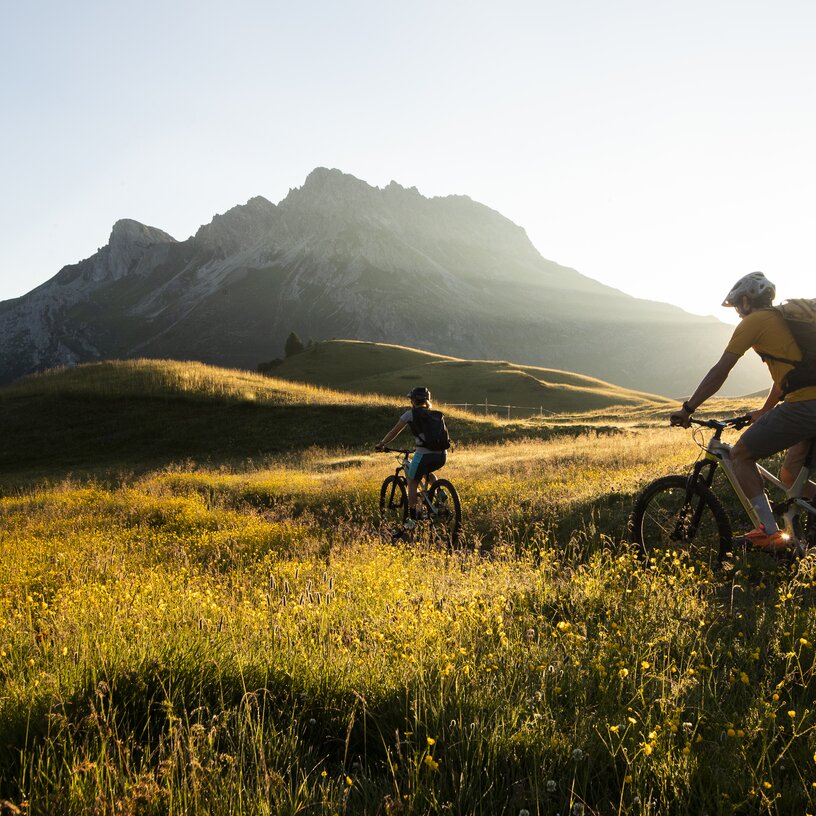 Zwei Radfahrer in einer bergigen Landschaft bei Sonnenuntergang.
