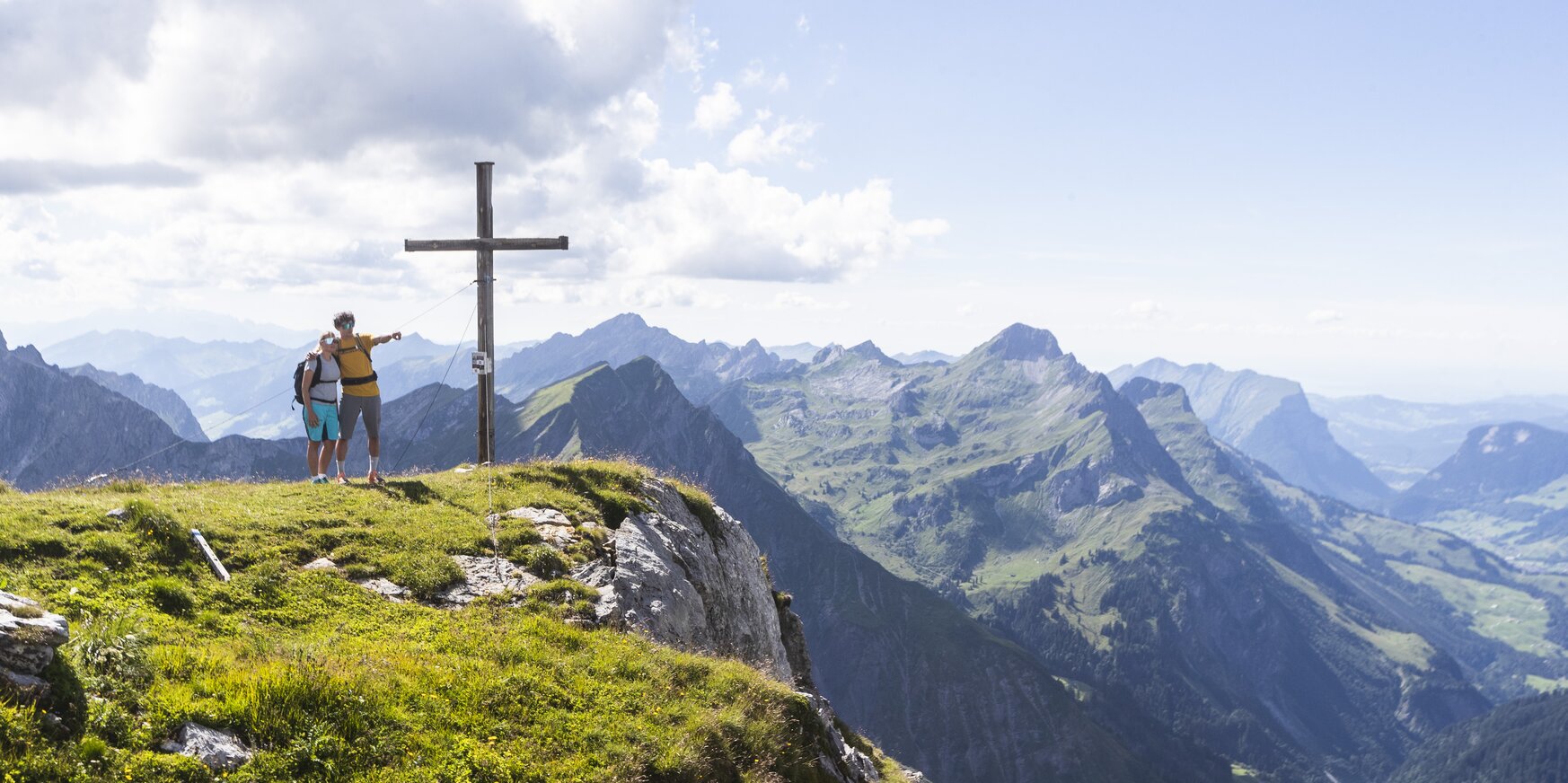Zwei Personen mit Wanderrucksäcken neben einem Gipfelkreuz mit Blick auf das malerische Bergpanorama.