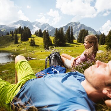 Ein Paar liegt entspannt in der Wiese neben einem Bergsee mit Alpenpanorama.