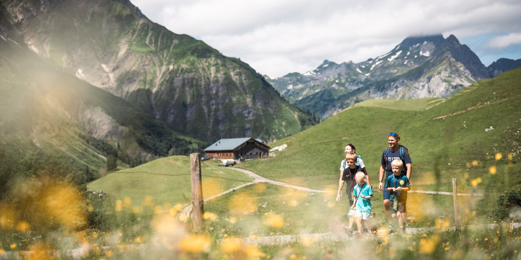 Familie wandert entlang eines Pfades über blühende Almwiesen, im Hintergrund ist eine Hütte zu sehen.