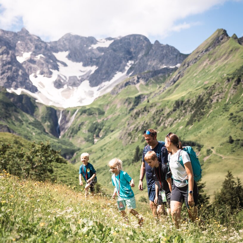 Eine Familie wandert auf einer blühenden Wiese mit Bergen im Hintergrund.