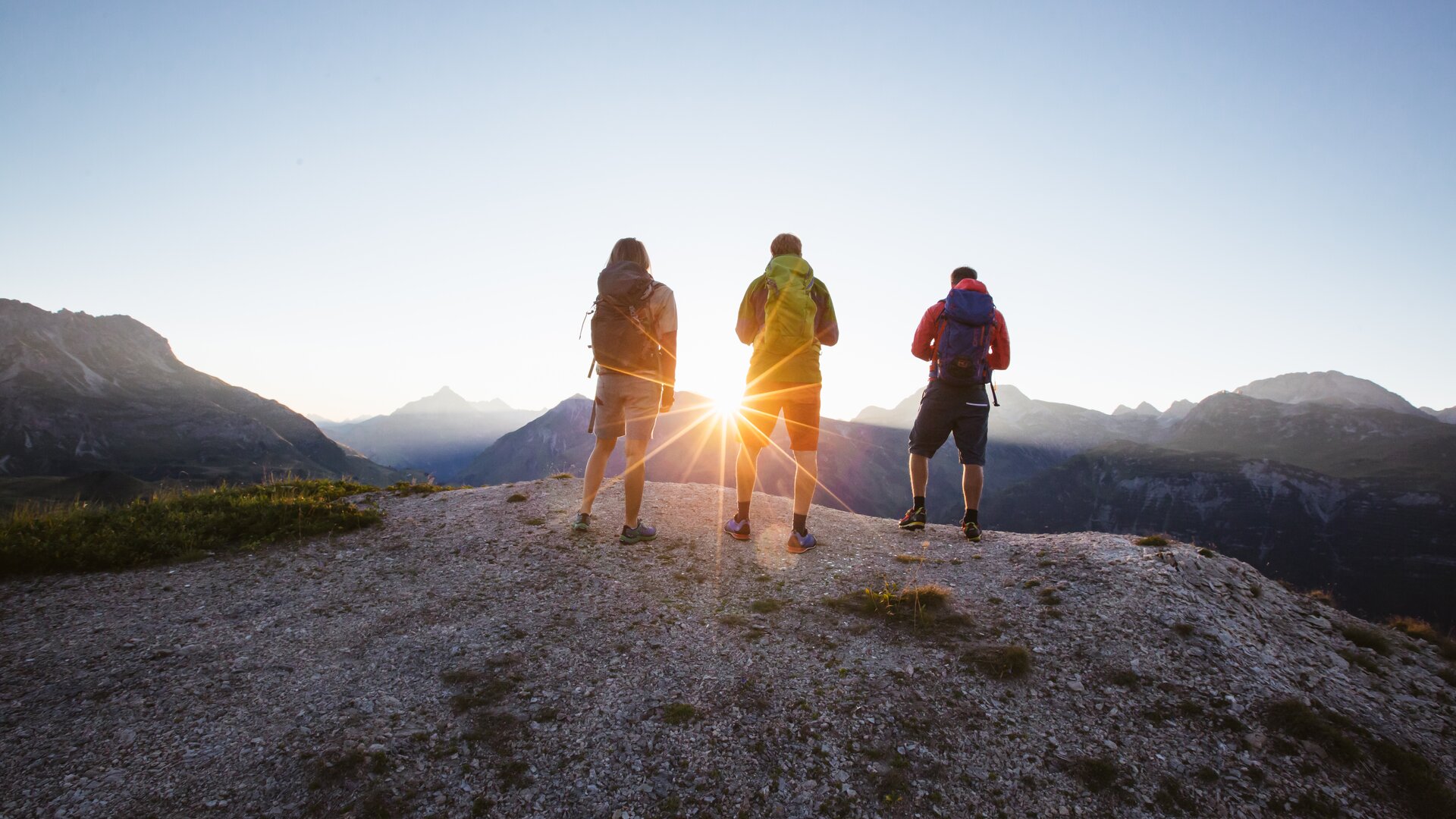 Drei Wanderer stehen auf einem Berggipfel und schauen in den Sonnenaufgang.