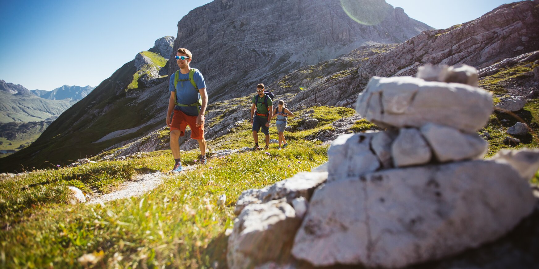 Eine Gruppe von Menschen wandert auf einem Bergpfad bei Sonnenschein, vorbei an einer Steinformation.