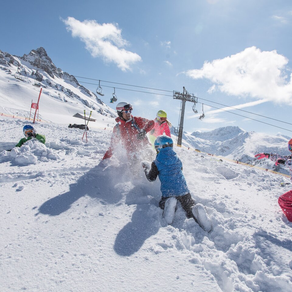 Kinder spielen im Schnee mit einem Erwachsenen, im Hintergrund ist ein Skilift zu sehen.