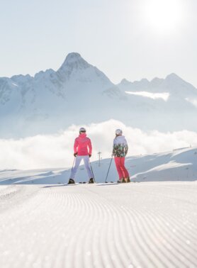 Zwei Skifahrer auf einer präparierten Skipiste vor schneebedeckter Bergkulisse.